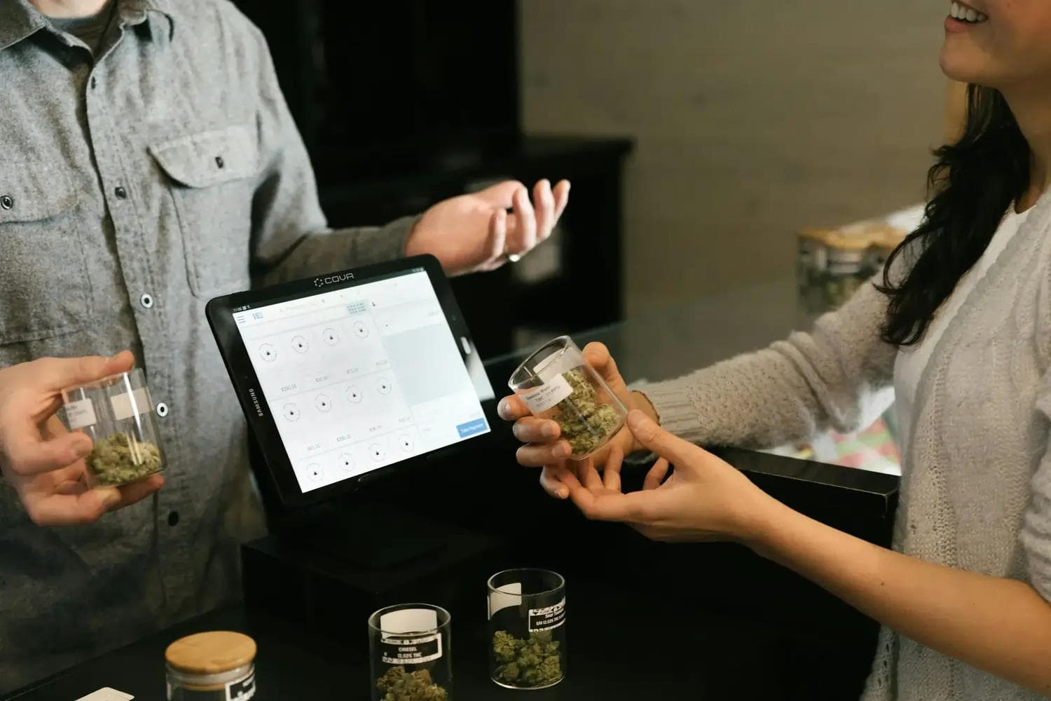 Hände tauschen Glas mit Cannabisblüten vor Tablet-Kasse