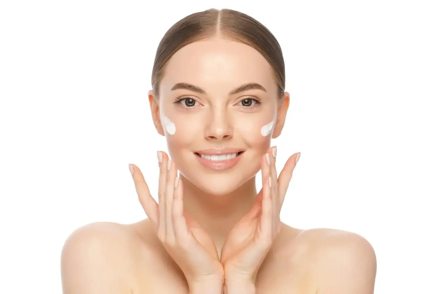 Junge Frau trägt Feuchtigkeitscreme auf Wangen, Hände am Gesicht, weißer Hintergrund