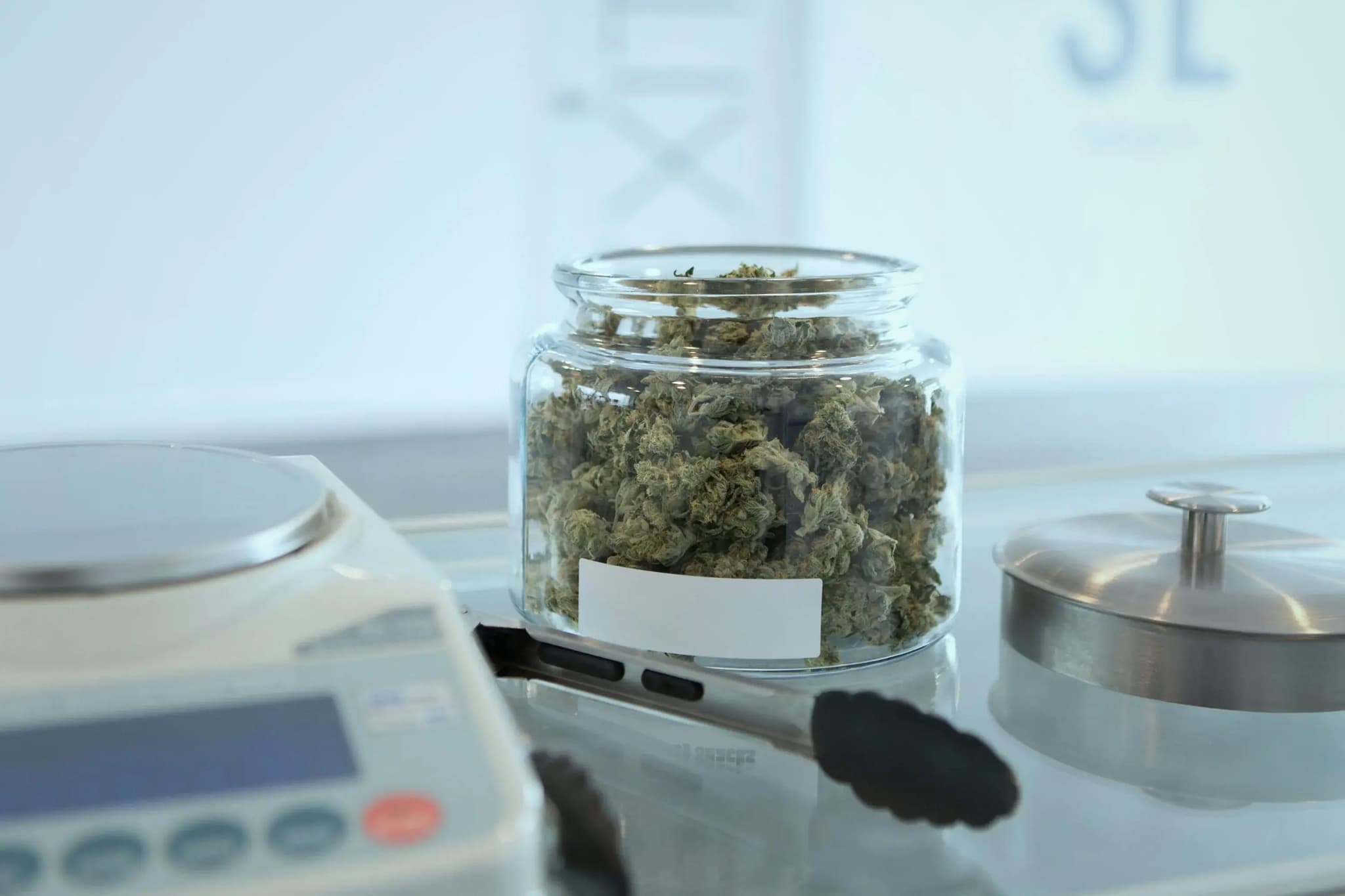 Glas mit getrockneten Cannabisblüten neben Waage und Zange