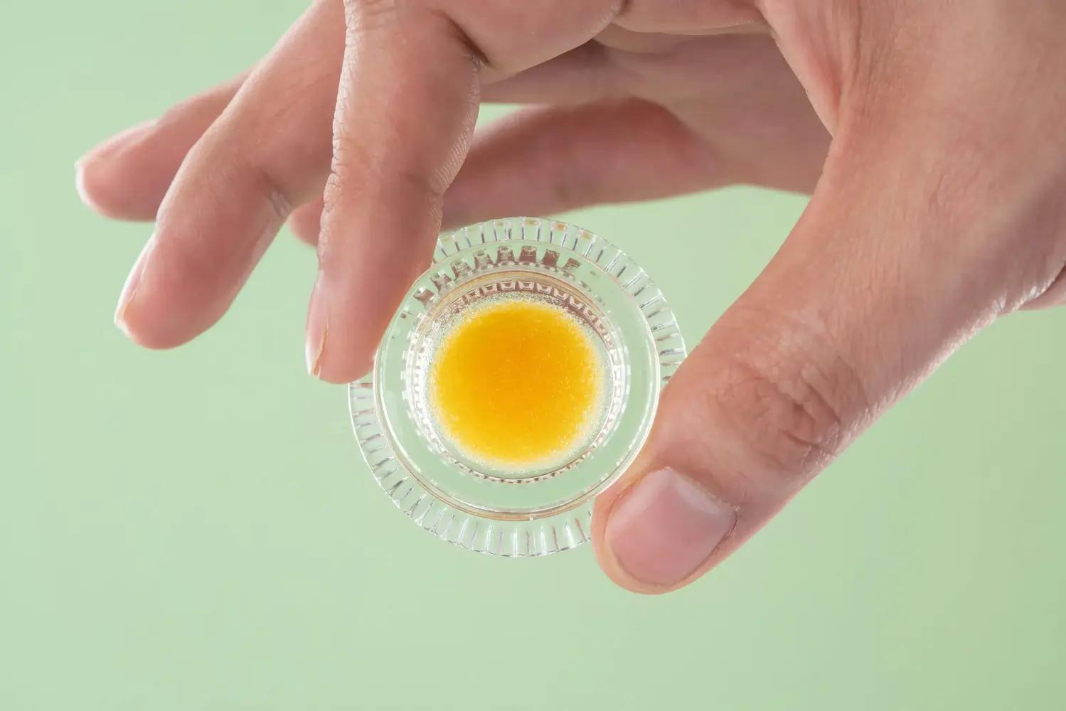 Hand hält Glas mit gelbem Cannabiskonzentrat vor mintgrünem Hintergrund