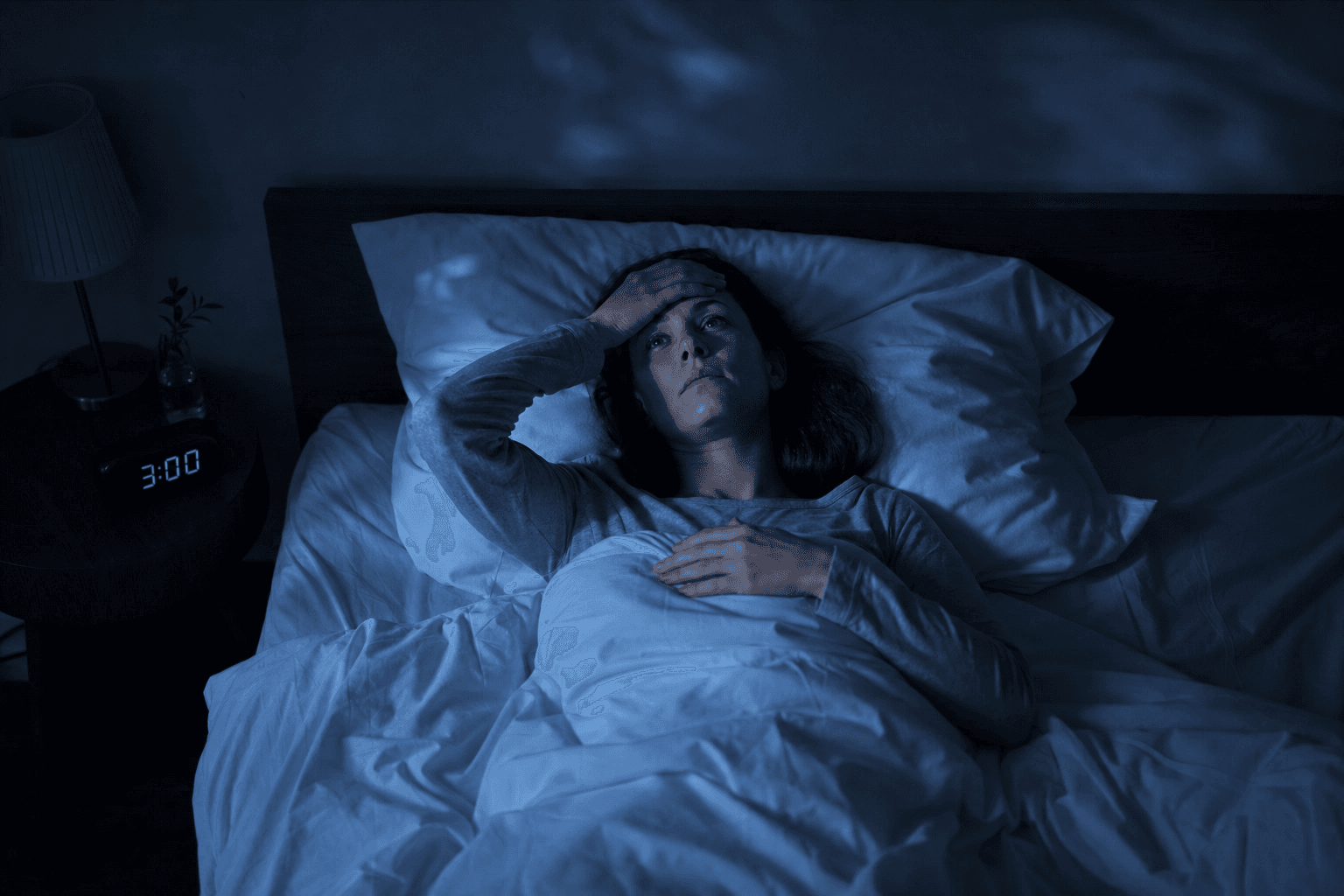Frau liegt nachts wach im Bett mit Hand auf der Stirn, Digitaluhr zeigt 3:00 Uhr, dunkles Schlafzimmer – Schlaflosigkeit durch Stress und innere Unruhe